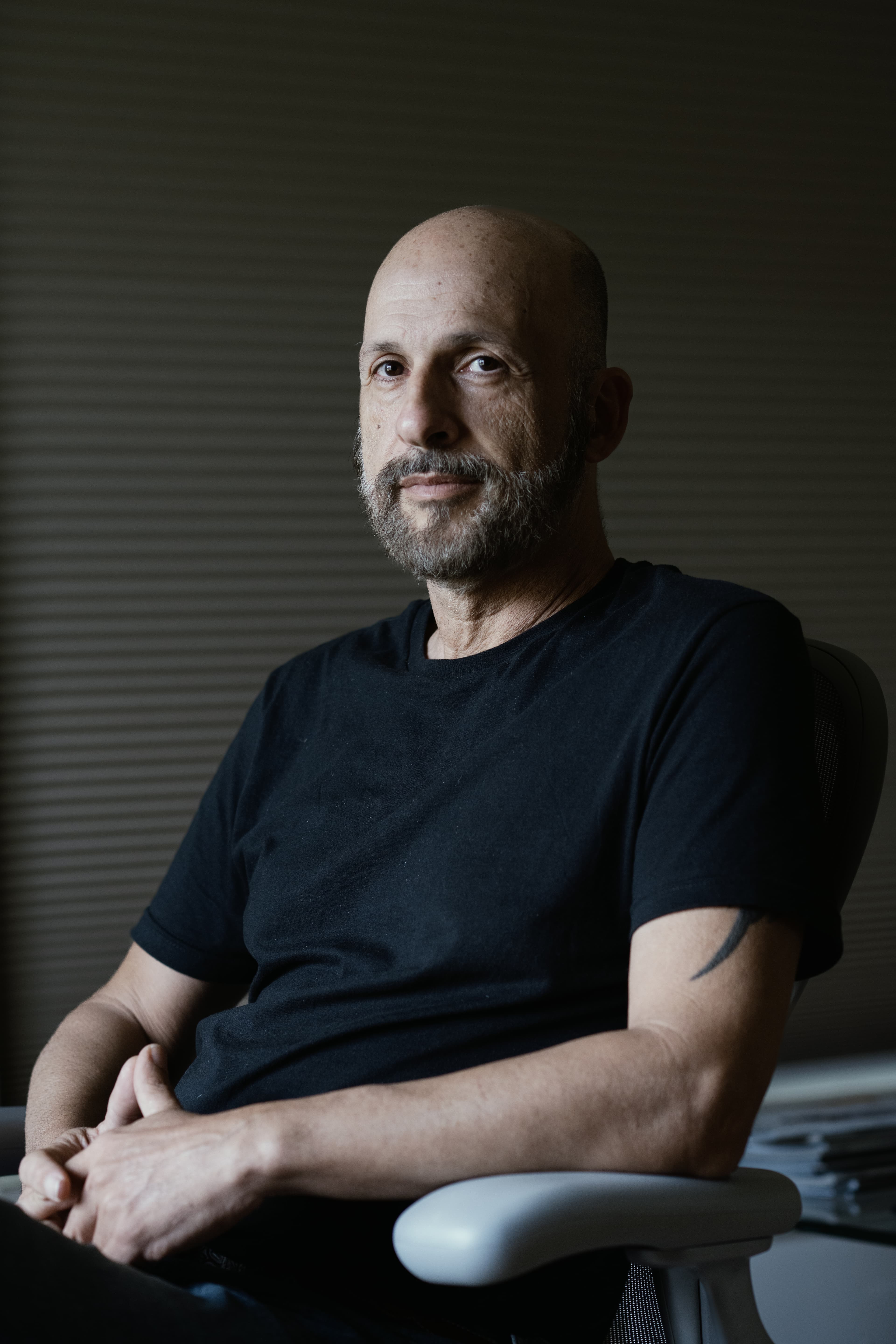Bald man with a grey beard sits in a chair against a dark slatted wall.